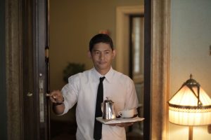 Positive ethnic male employee carrying tray with coffee while serving guest in cozy hotel room